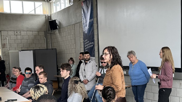 Die Schulleitung, zwei Lehrerinnen sowie Referentinnen und Referenten sitzen bzw. stehen vorne in der Aula.