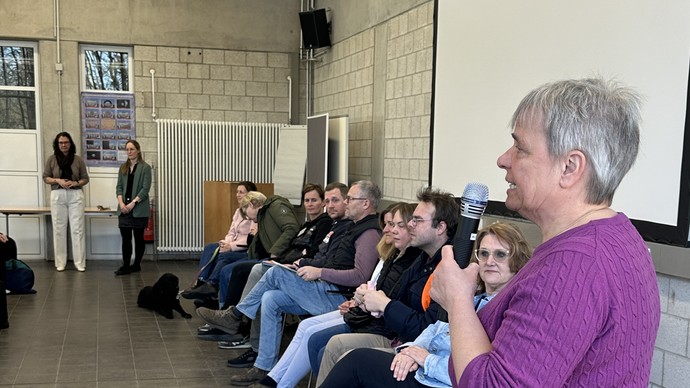 Lehrerin am Mikrophone steht neben sitzenden Referenten und Referentinnen