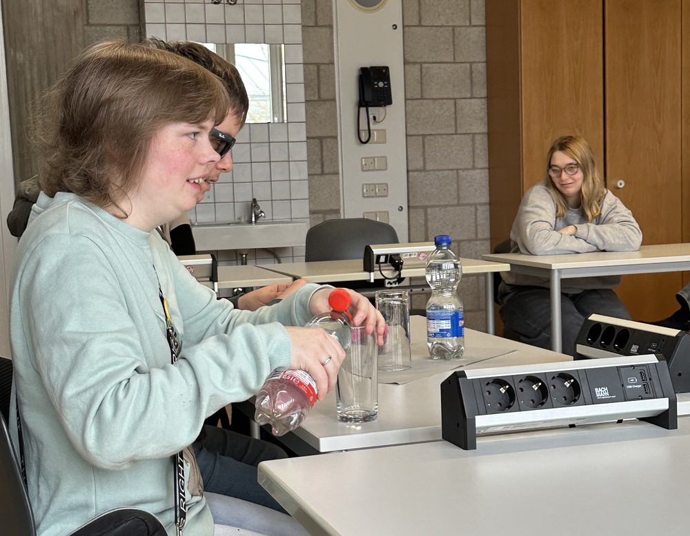 Die Referentin sitzt vorne, gießt sich Wasser ins Glas und Schülerin und Schüler hören ihr aufmerksam zu.