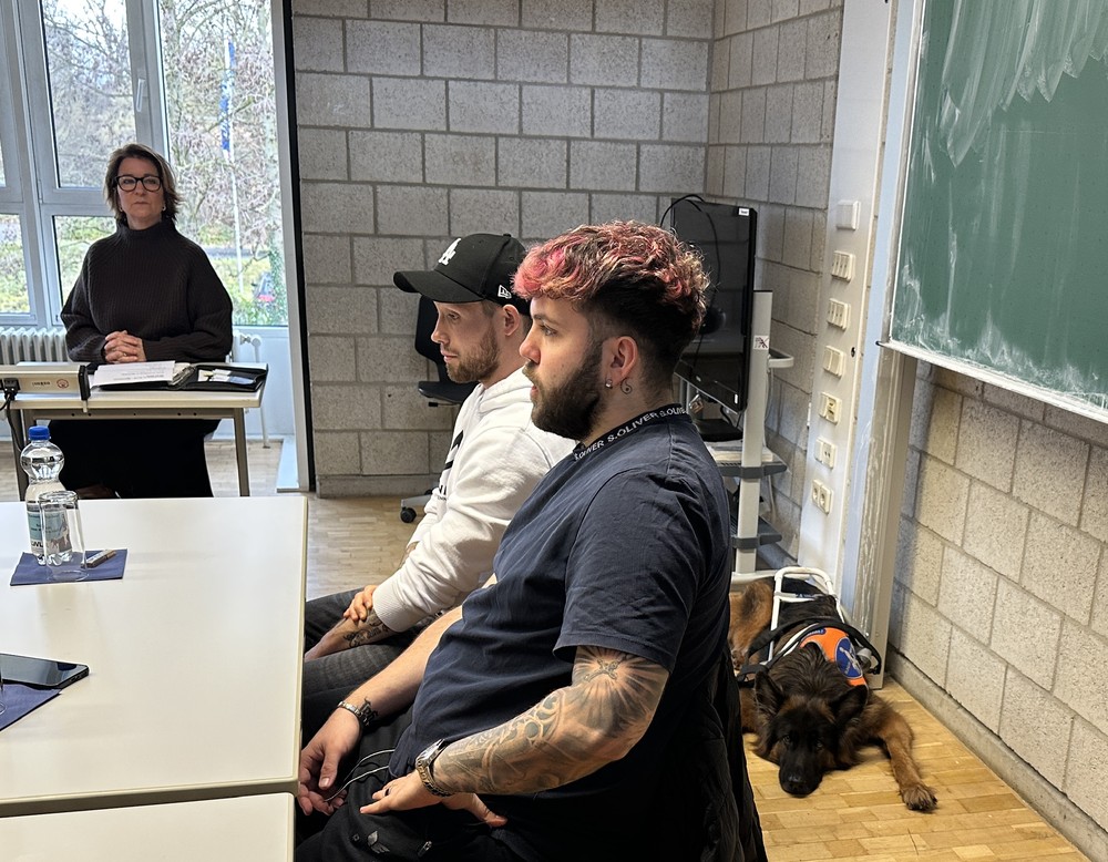 Die Referenten sitzen mit einem liegenden Blindenhund vor der Tafel und leiten den Workshop