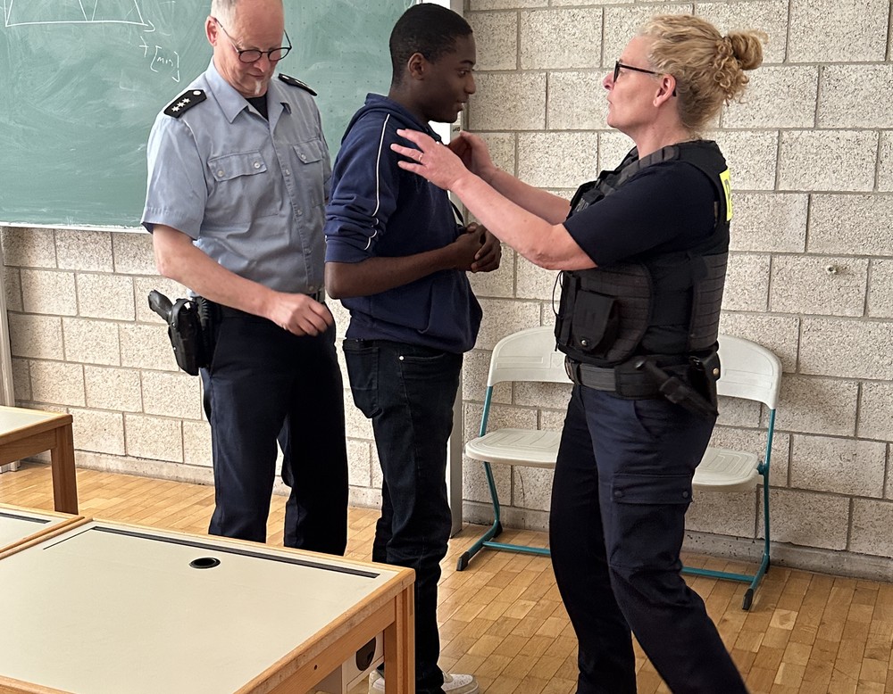 Ein Schüler steht, ein Polizist hinter ihm und eine Polizistin vor ihm. Sie zeigt, wie ein Handy aus seiner Tasche genommen wird.