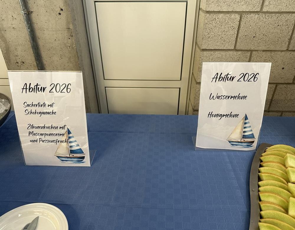 Zwei Schilder auf einem Buffet informieren über die angebotenen Kuchen- und Obstsorten. Rechts daneben liegt eine Platte mit Melonenscheiben.