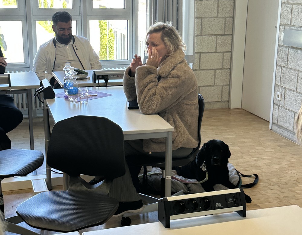 Die Referentin sitzt und hält ihren Vortrag. Unter ihr liegt ihr Begleithund. Rechts von ihr sitzt ein Schüler und hört zu.