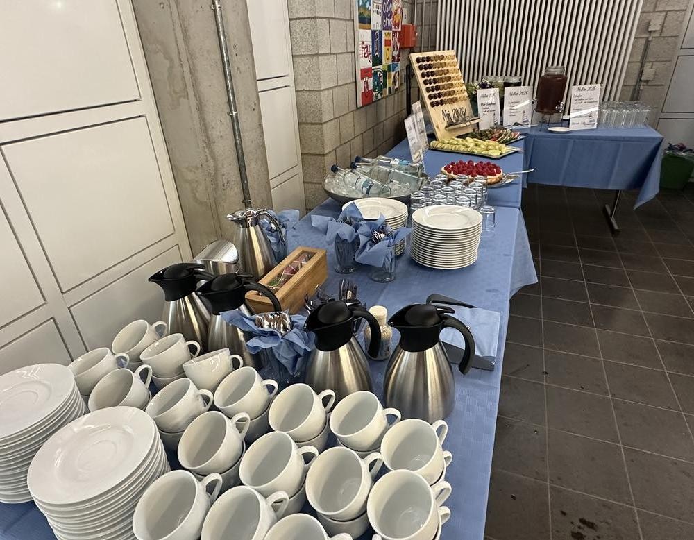 Ein Buffet mit Kaffee, Wasser, Säften, Obst und Kuchen ist aufgebaut. Daneben stehen Tassen, Teller, Gläser und Besteck bereit.