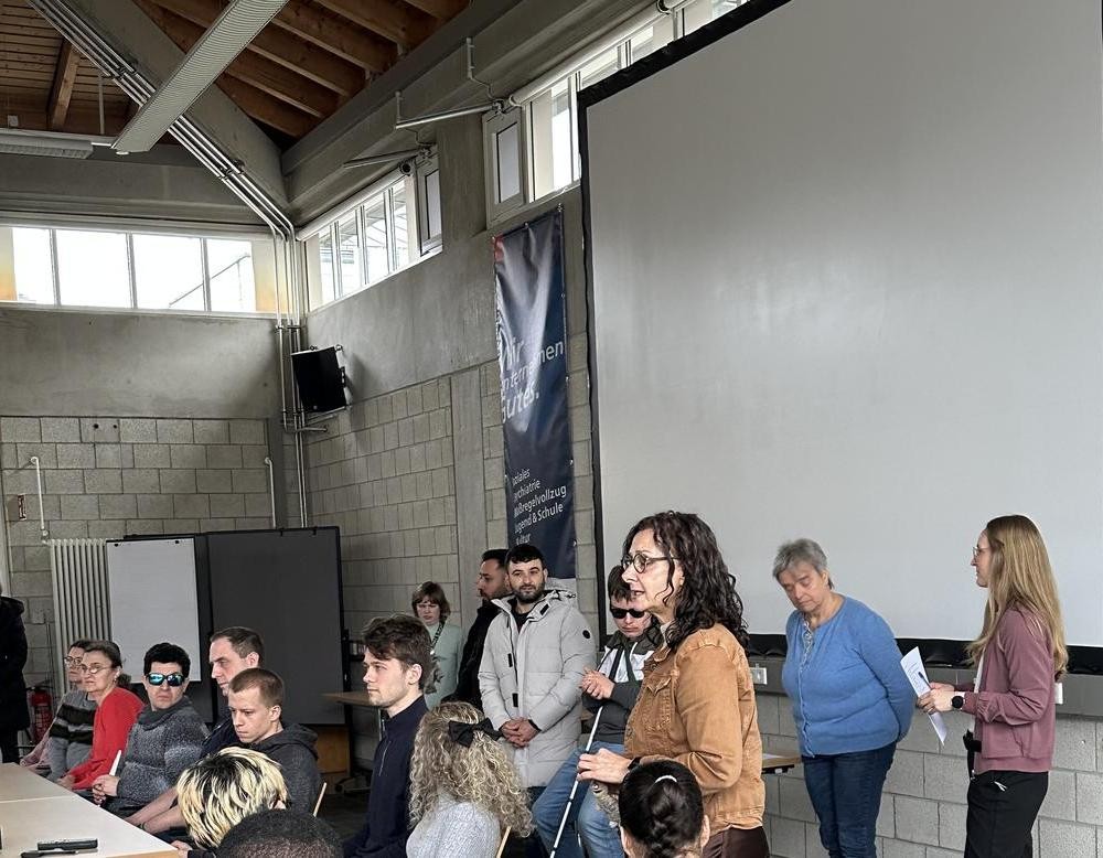 Die Schulleitung, zwei Lehrerinnen sowie Referentinnen und Referenten sitzen bzw. stehen vorne in der Aula.