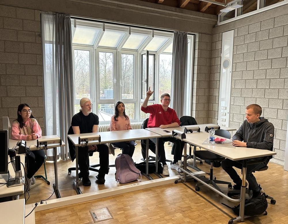 Auf der linken Seite sitzen zwei Schülerinnen und zwei Schüler. Einer der Schüler meldet sich, während vorne der Referent sitzt.