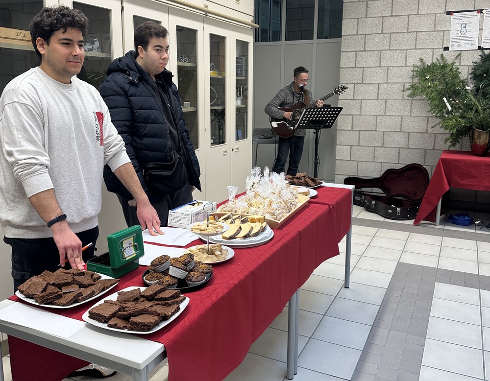 Zwei Schülerinnen bzw. Schüler stehen hinter einem Tisch mit Kuchen und Gebäck, daneben der Schulsozialarbeiter mit einer Gitarre.
