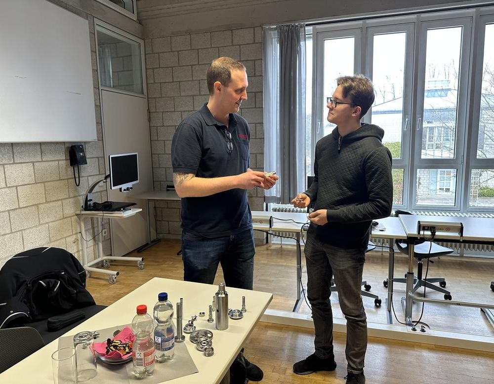 Der Referent steht einem stehenden Schüler gegenüber. Beide lächeln und halten Metallstücke in der Hand.